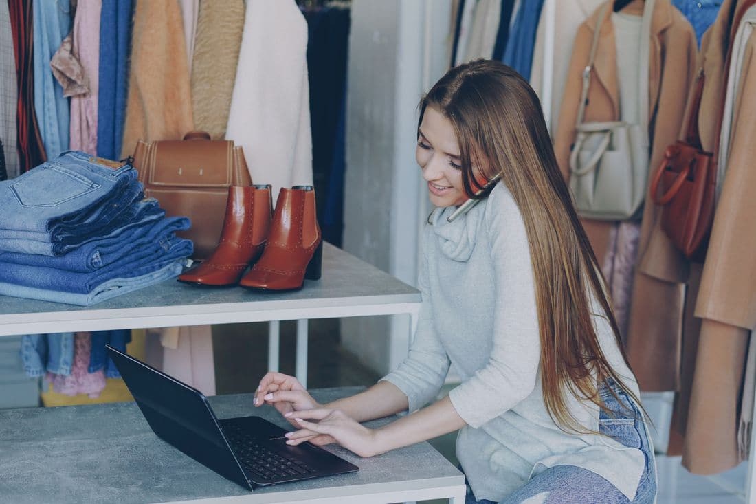 A woman with long hair talking on the phone while using a laptop at a table in a clothing shop