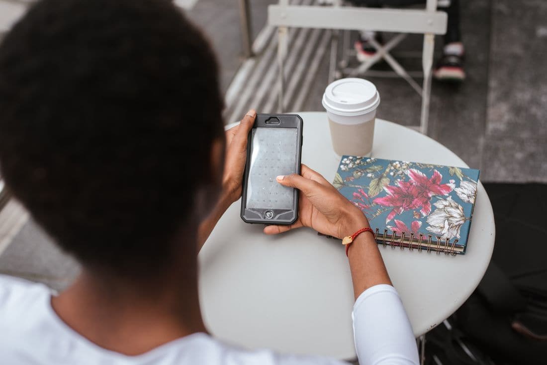 A person at a cafe table holding a phone with a calendar on the screen, beside a takeaway cup and a floral notebook