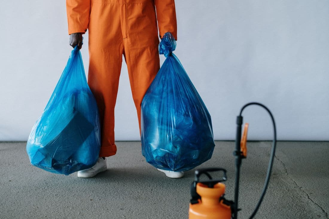 A worker in orange overalls carrying two full rubbish bags beside a spray bottle