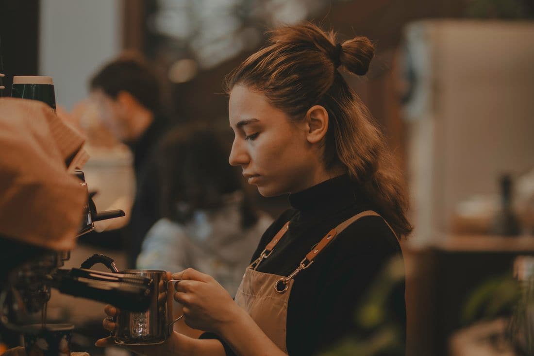 A barista in a dark apron steaming milk at an espresso machine, with coworkers blurred in the background