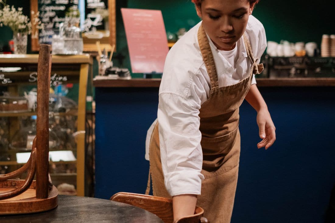 A cafe worker in a white shirt and apron putting chairs up on a table in front of the counter