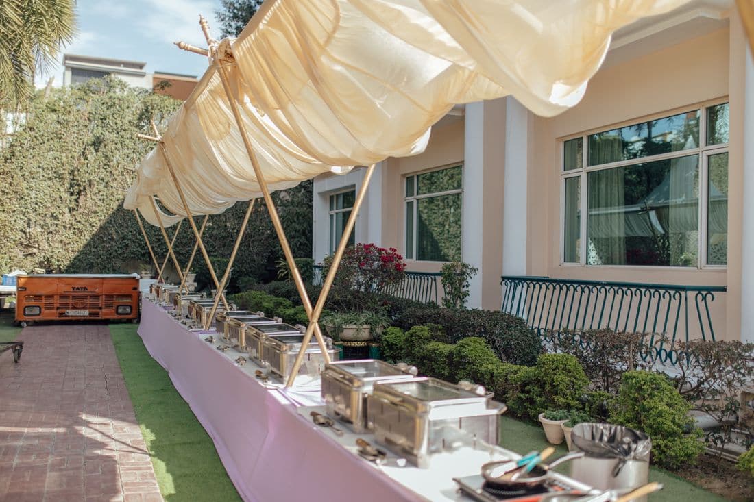 A long outdoor buffet table of chafing dishes set up under a fabric canopy