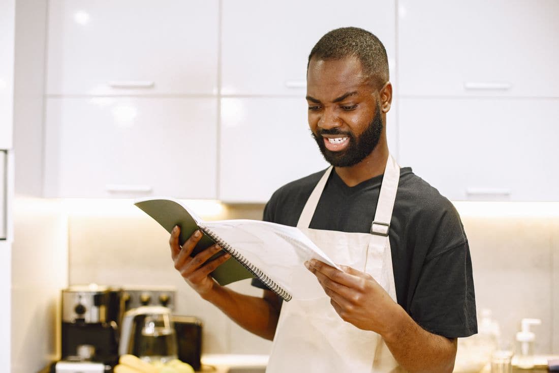 A man in an apron frowning as he reads a notebook in a kitchen