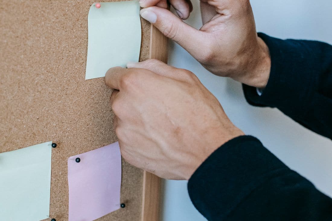 Two hands pressing a pin into a pale green note on a cork board, beside a pink note and another green note