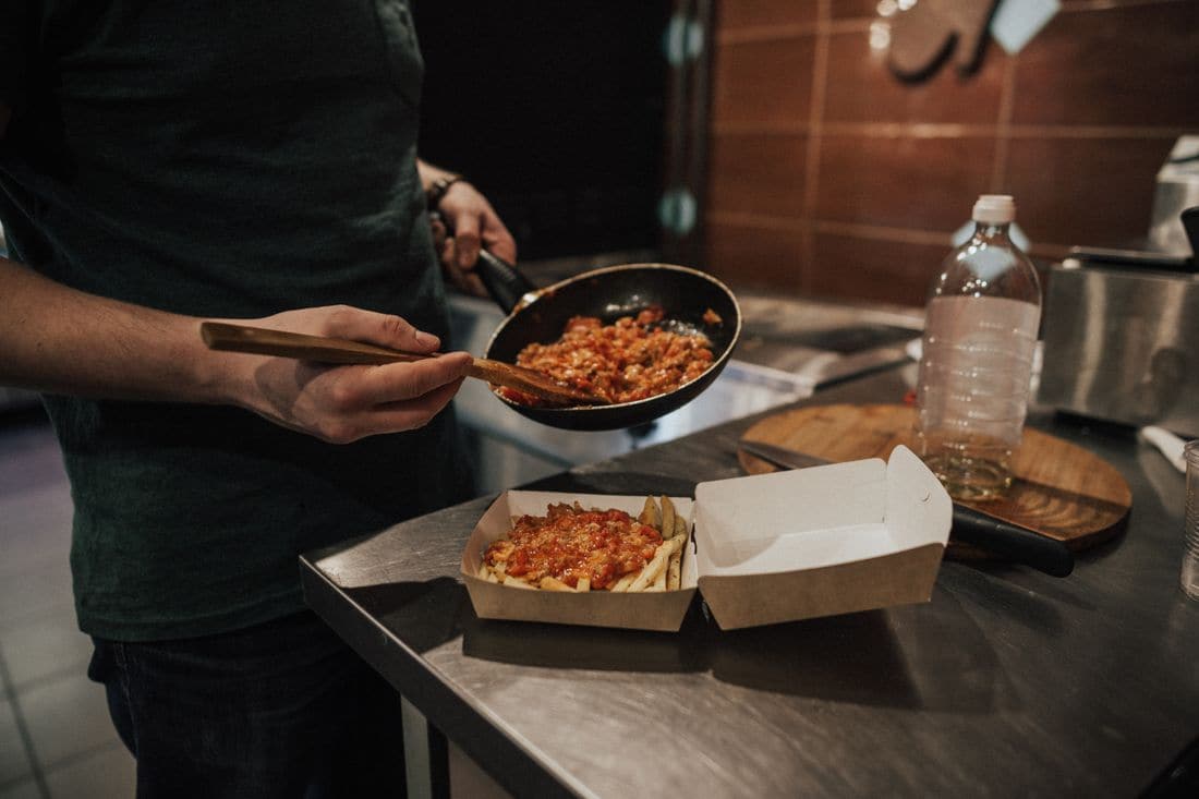 A cook spooning pasta from a pan into a takeaway box on a steel kitchen bench