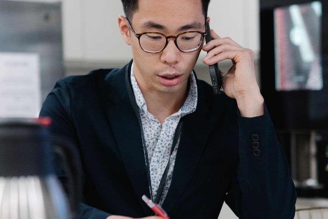 A man in glasses and a dark blazer talking on a mobile phone while writing notes with a red pen