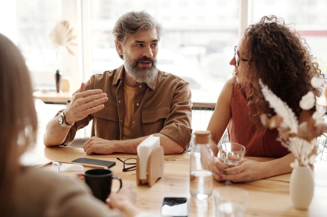 Three people talking around a wooden table with glasses of water in a bright cafe
