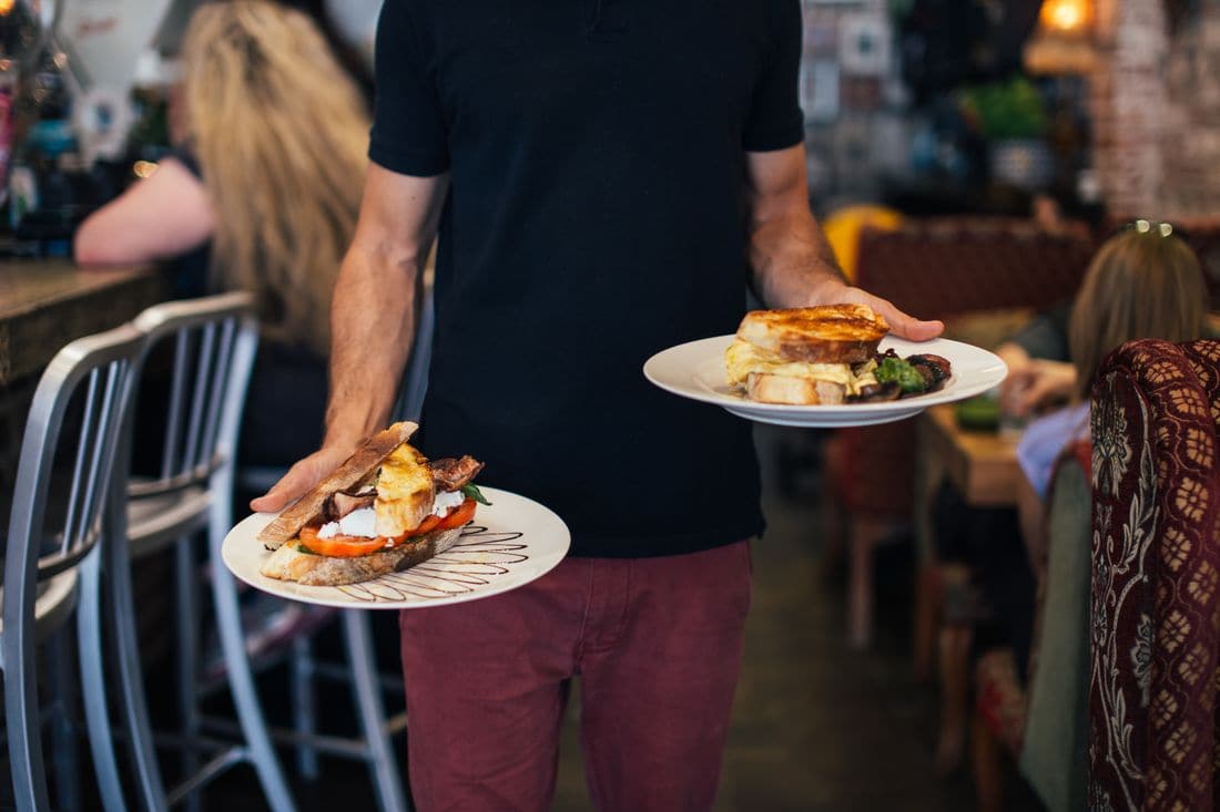 A waiter in a black shirt carrying two plates of breakfast dishes past diners in a cafe