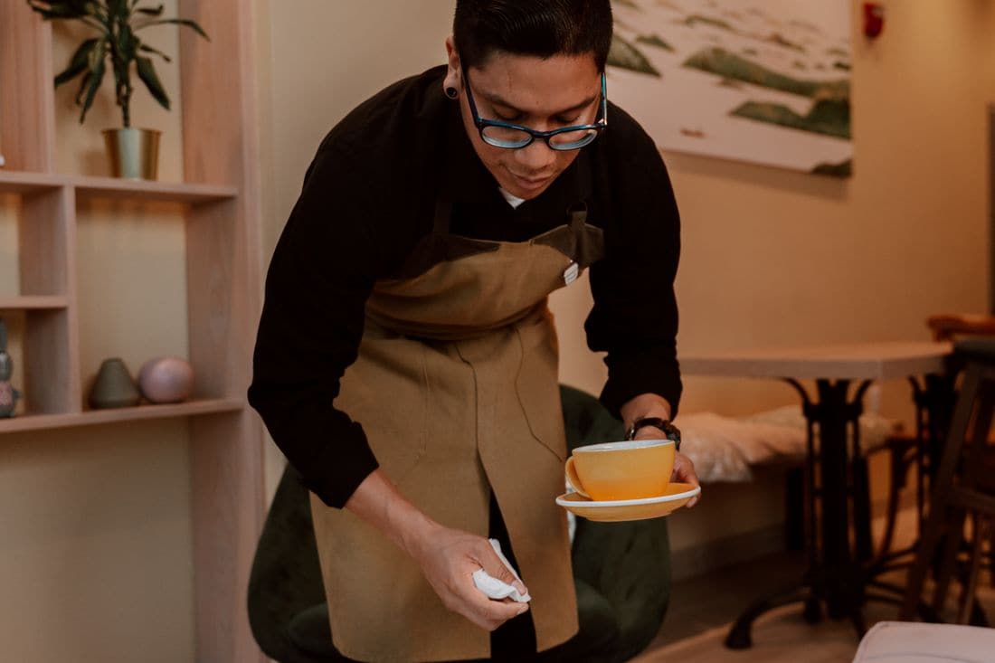 A cafe worker in an apron wiping a table while holding a yellow cup and saucer