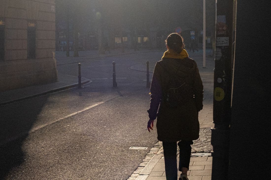 A person in a dark coat and yellow scarf walking along a city footpath in low early morning sun