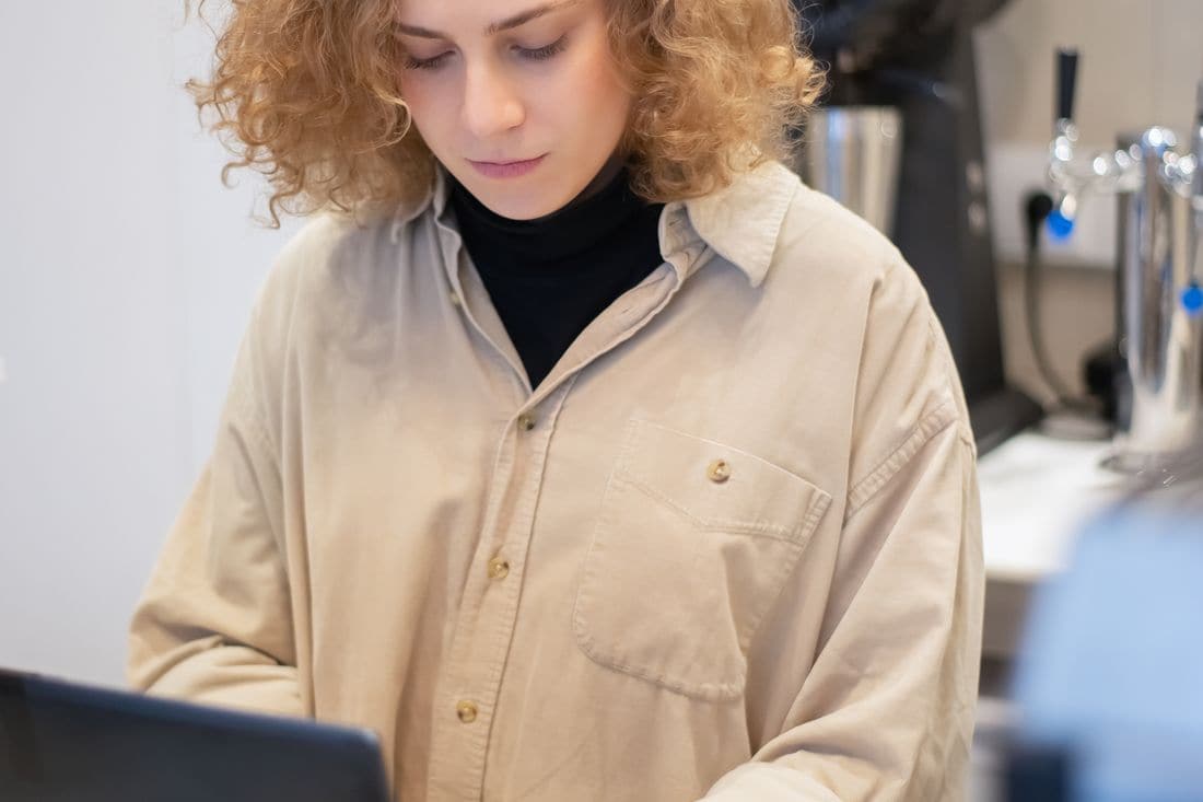 A woman in a beige shirt looking down at a laptop on a counter, with chrome taps behind her