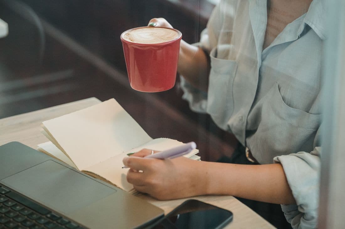A person writing in an open notebook at a table, holding a red cup of coffee, with a laptop and a phone beside the notebook