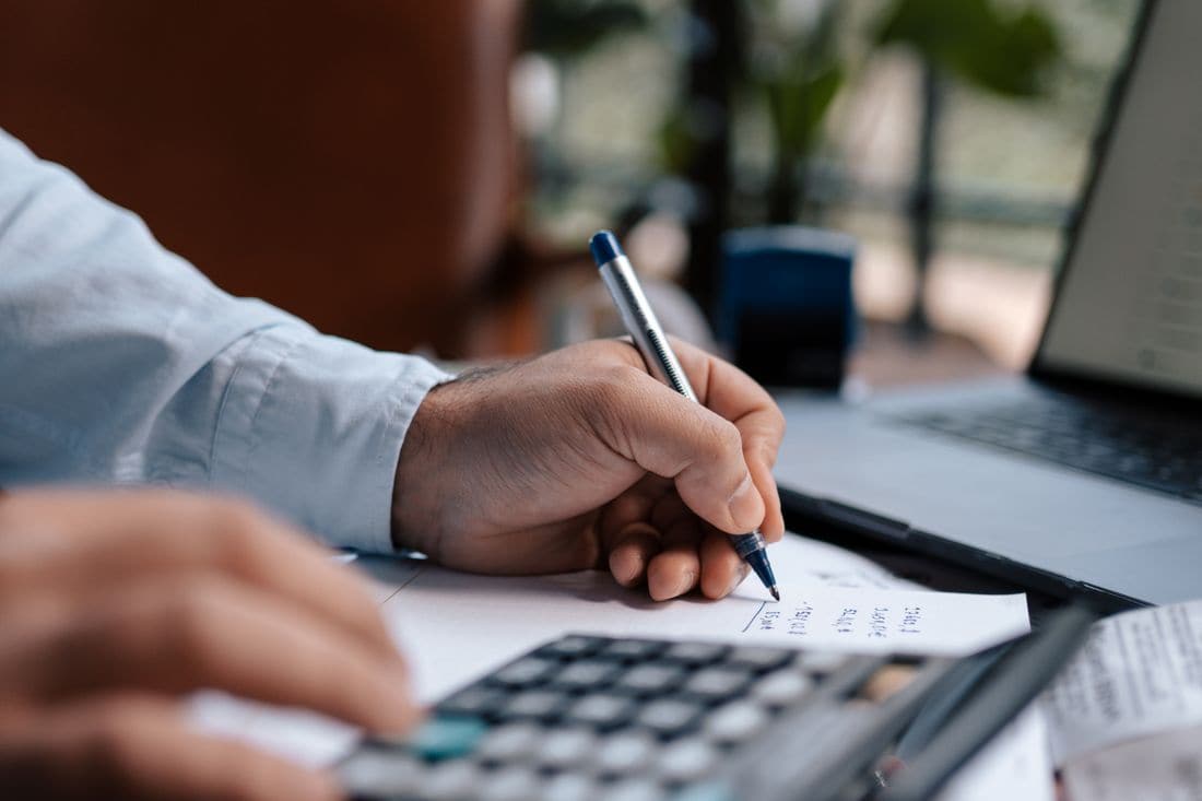 A man writing figures on a sheet of paper with a pen, beside a calculator and an open laptop