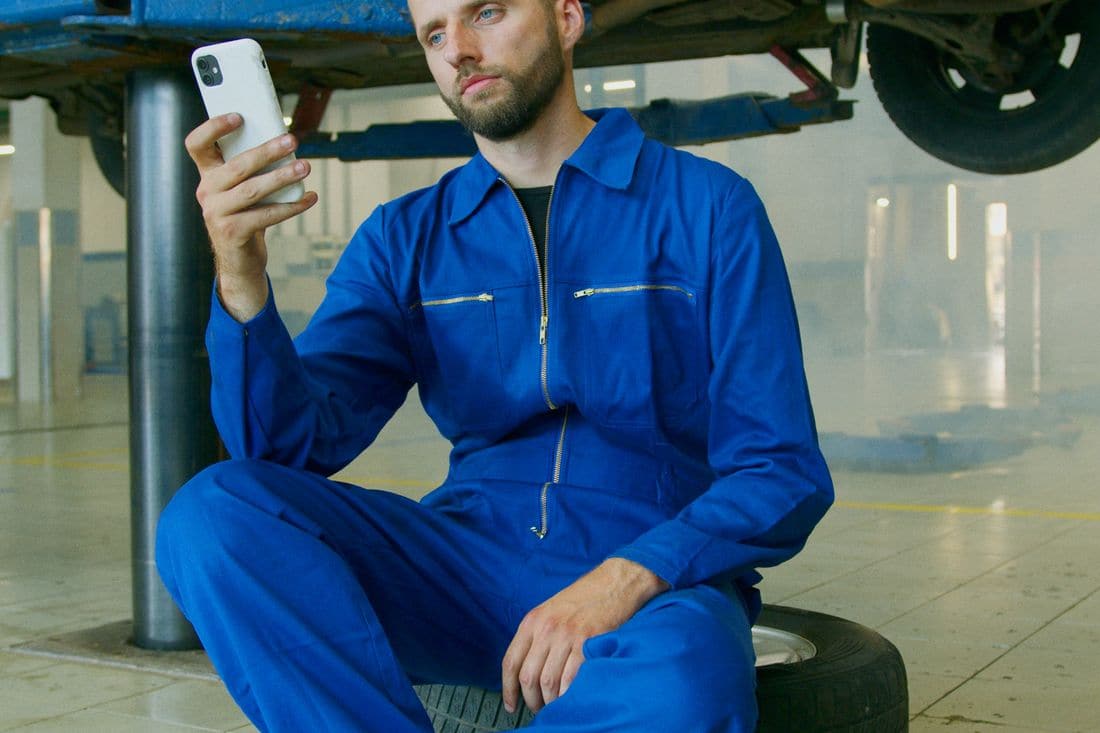 A mechanic in blue overalls sitting on a tyre under a raised car, looking at a phone in his hand