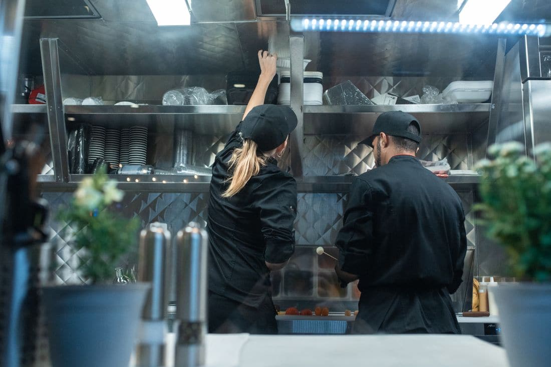 Two cooks in black caps working at the shelves of a small commercial kitchen