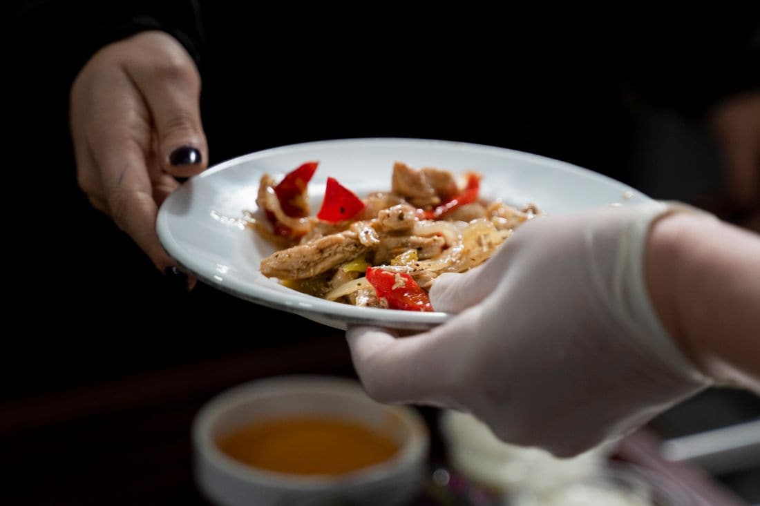 A gloved hand passing a plate of chicken and red capsicum to another person’s outstretched hand