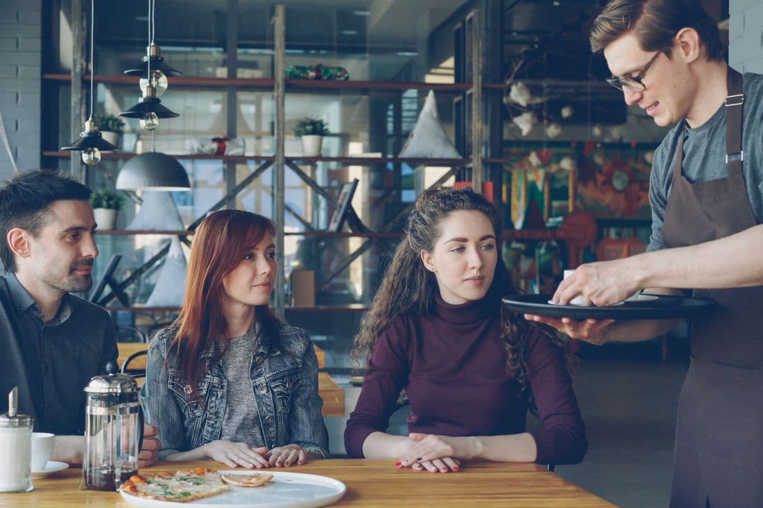 A waiter in a brown apron bringing a tray to three customers seated at a wooden table in a cafe