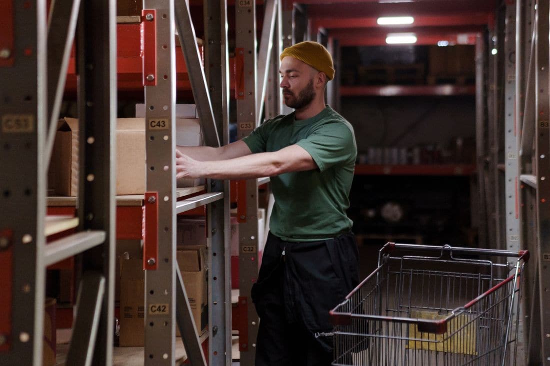 A worker in a beanie stacking boxes on stockroom shelves beside a trolley