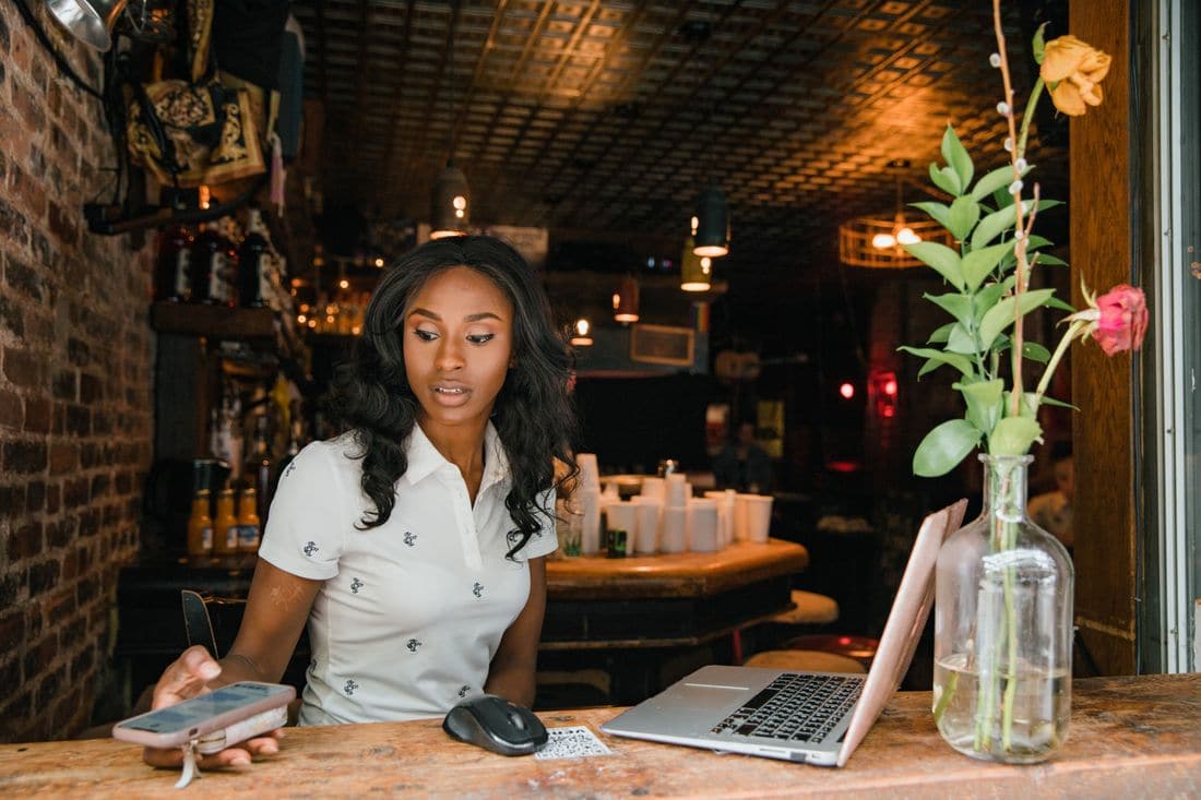 A woman at a bar counter checking her phone beside an open laptop and a vase of flowers