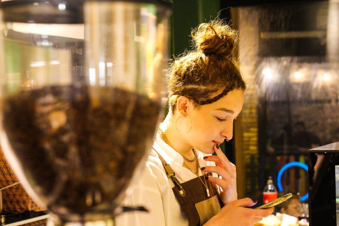 A barista in a brown apron looking at a phone behind a coffee grinder in a dimly lit cafe