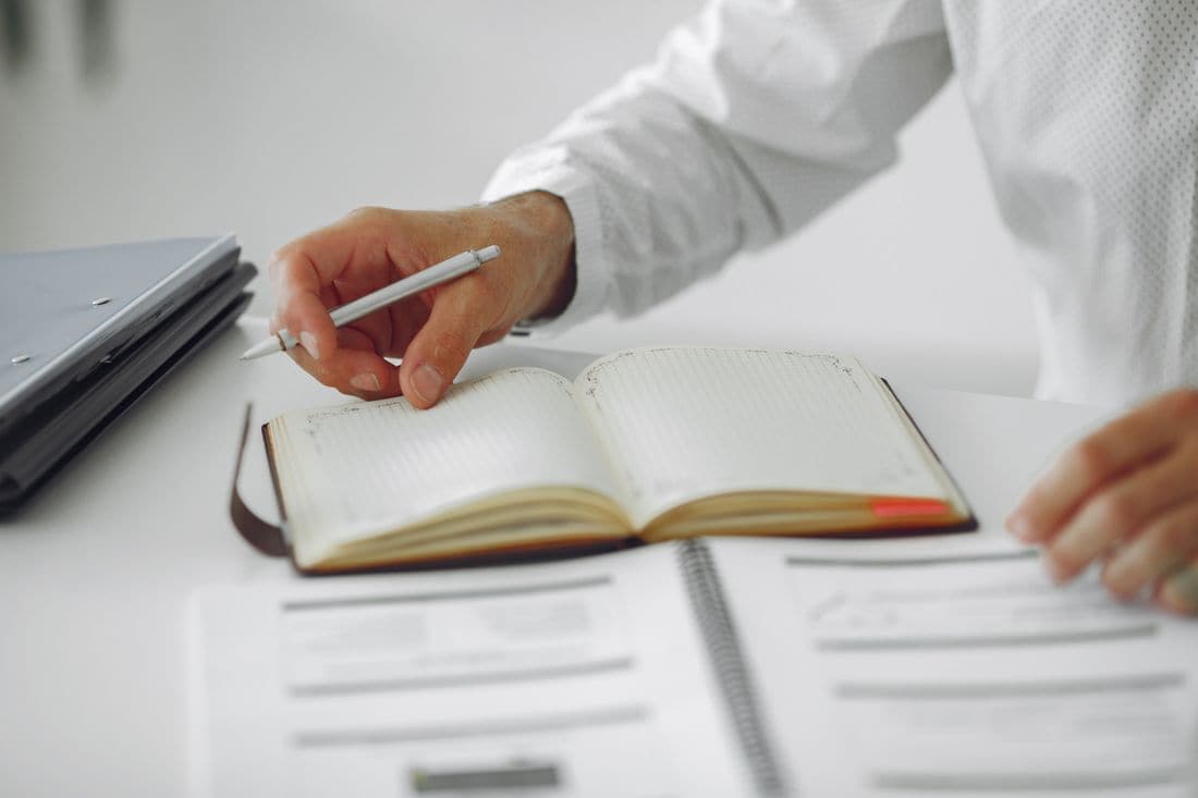 A man writing in a diary at a desk beside a laptop and printed papers