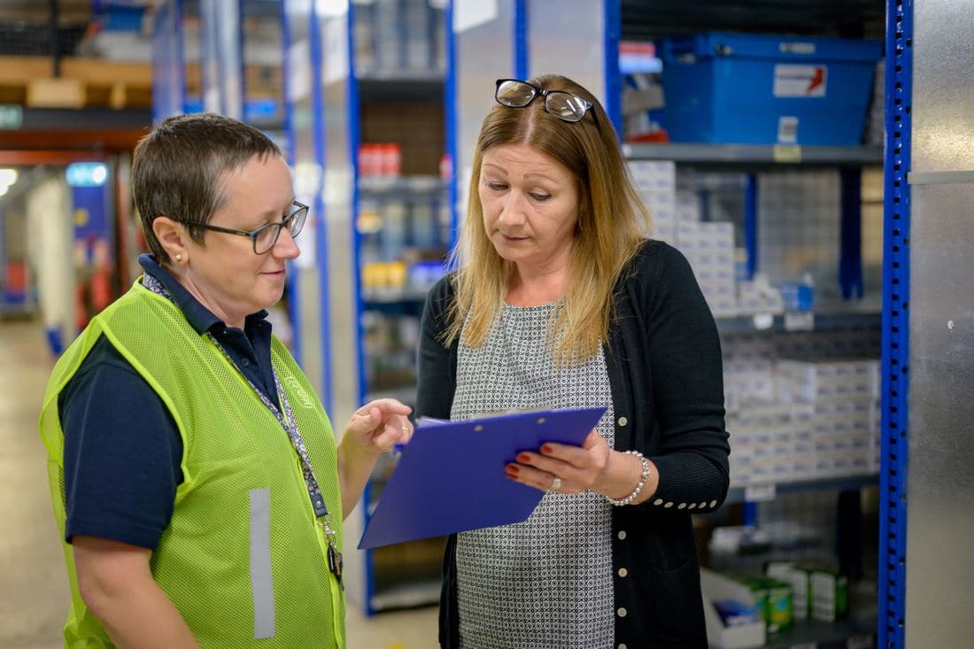 Two women in a warehouse aisle going over a list on a blue clipboard
