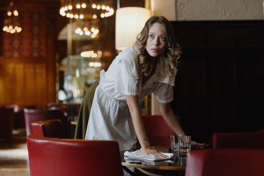 A waitress in a white uniform wiping a table with a napkin in a dimly lit restaurant with red leather chairs