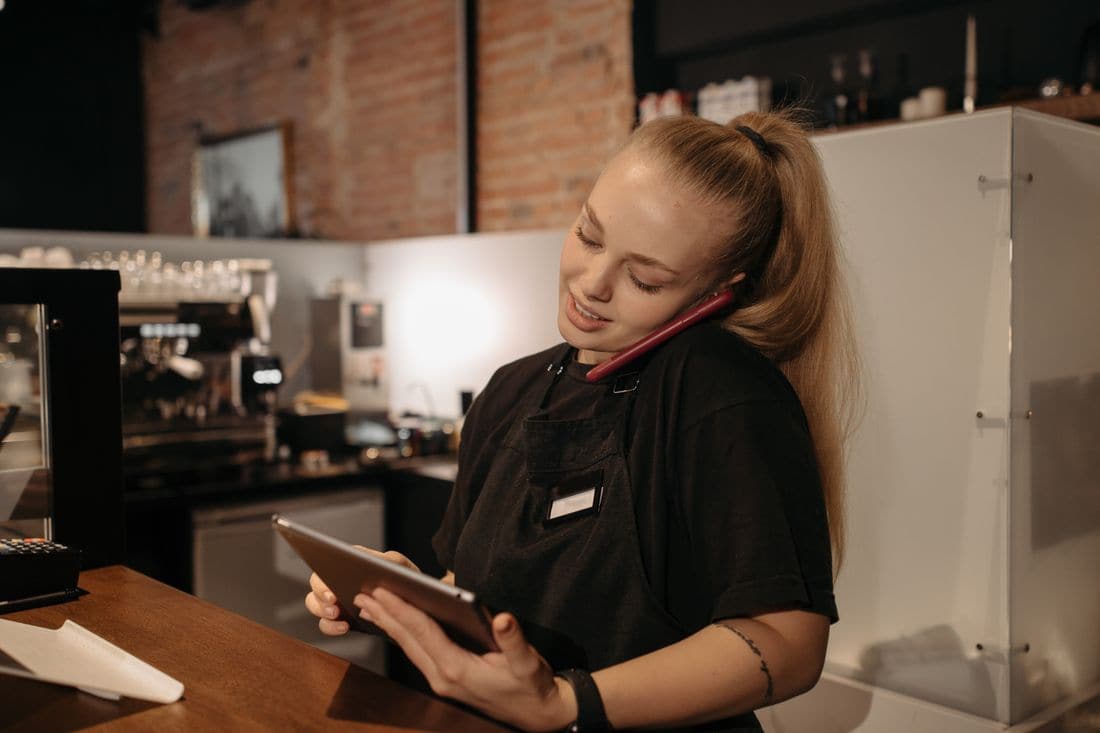A cafe worker in a black apron talking on a phone while holding a tablet at the counter