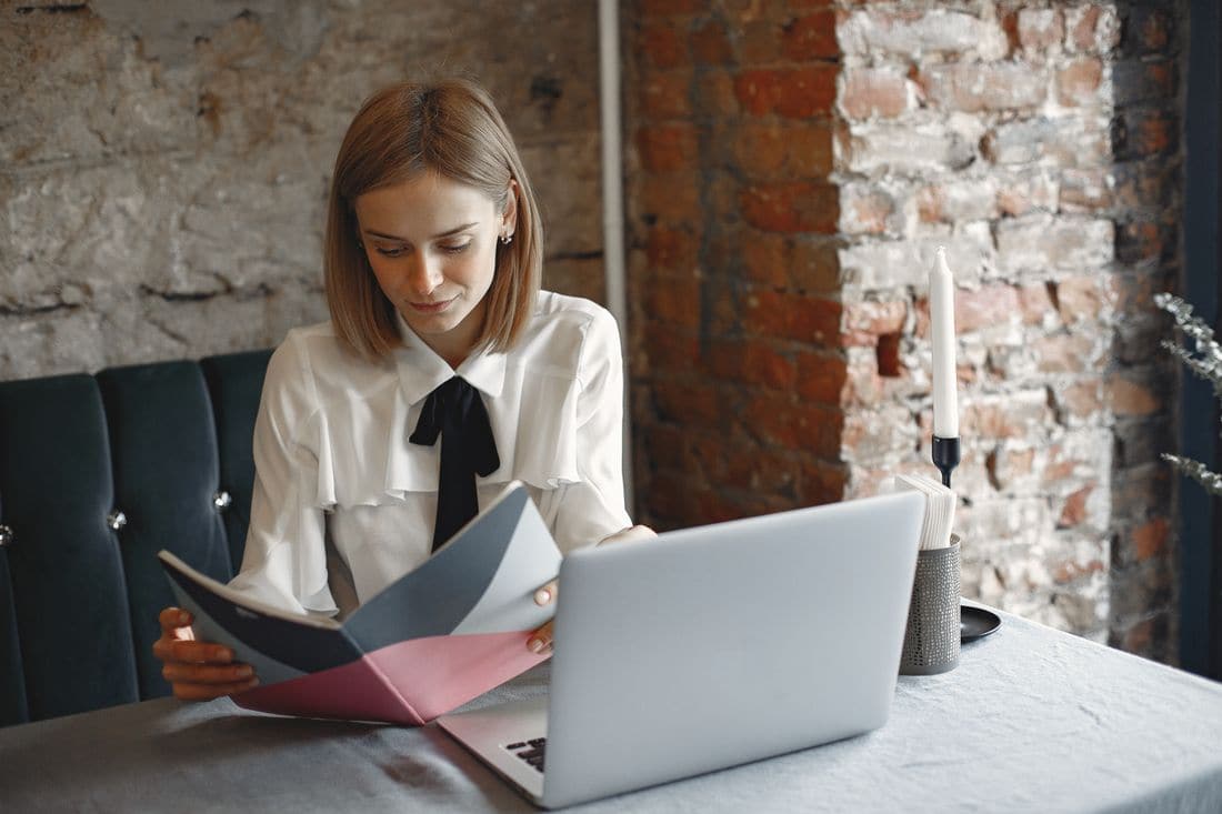 A woman at a restaurant table reading a folder beside an open laptop