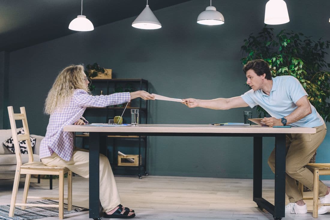 A man and a woman at opposite ends of a long table both holding the same sheet of paper