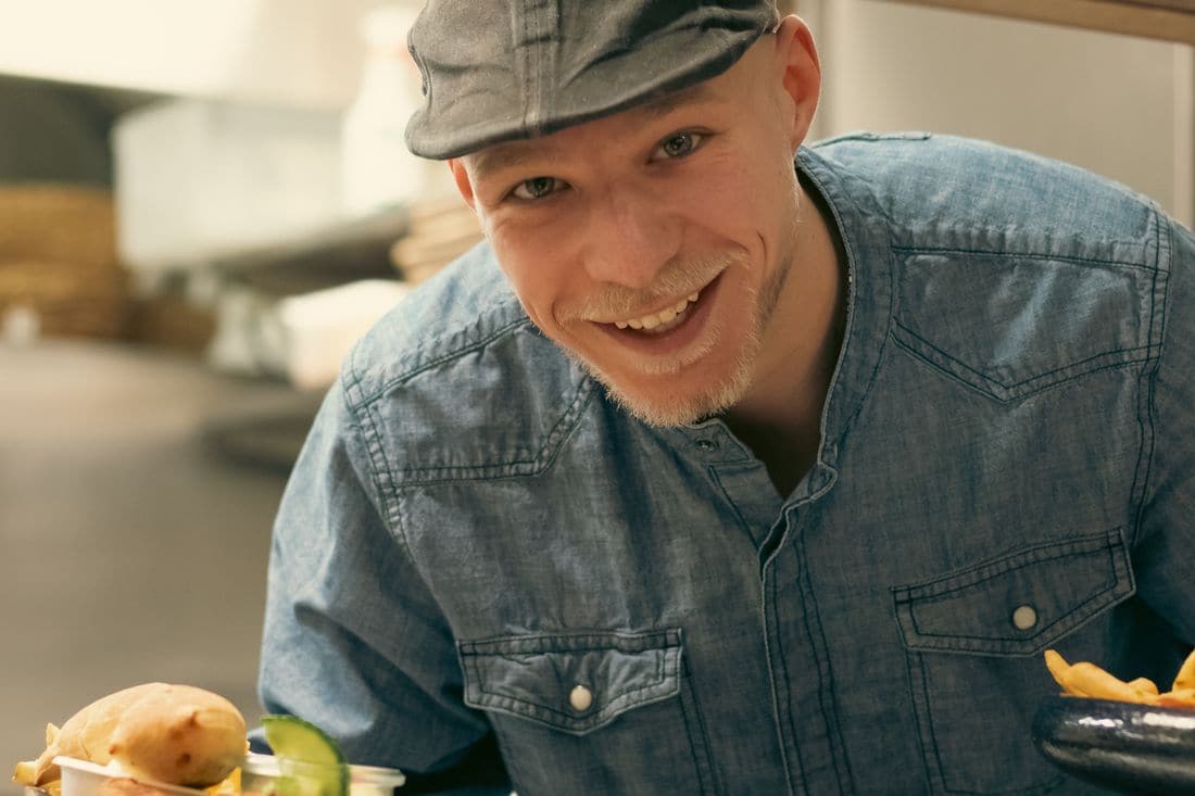 A smiling cook in a flat cap and denim shirt leaning over a counter beside a burger and a bowl of chips