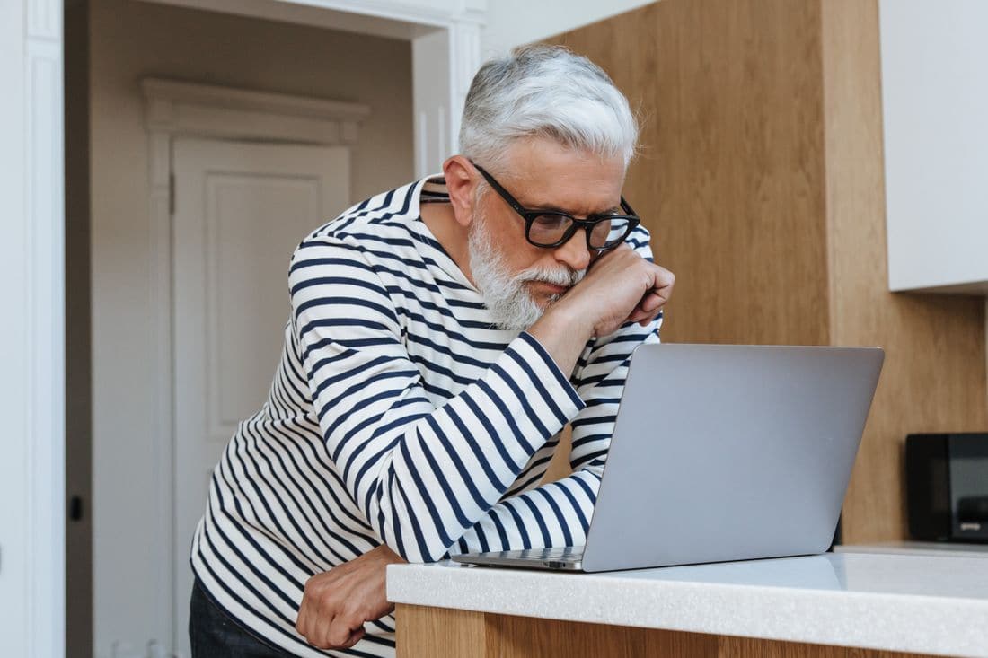 An older man in glasses leaning over a laptop on a kitchen bench, reading closely