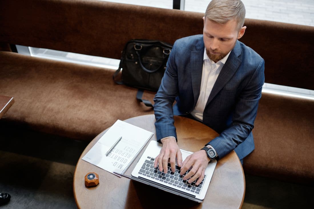 A man in a suit typing on a laptop at a round table beside a notebook
