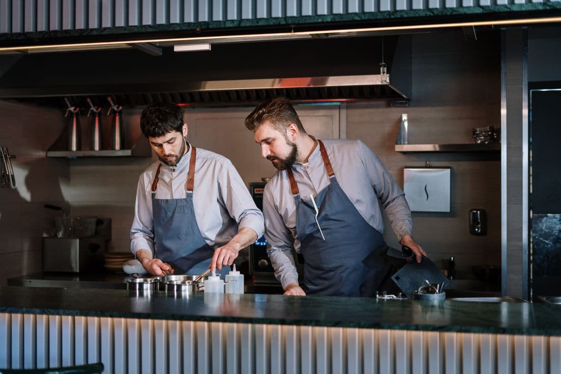 Two chefs in aprons preparing food side by side at an open kitchen counter