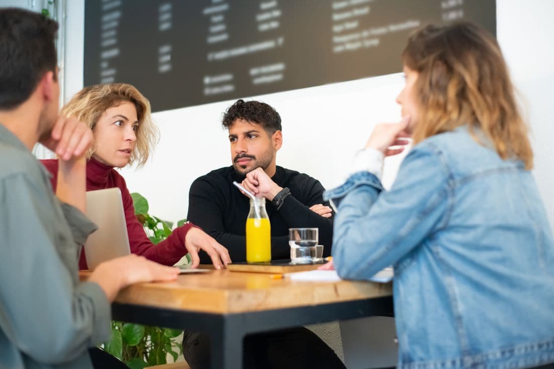 Four people talking around a cafe table with a laptop and a notebook