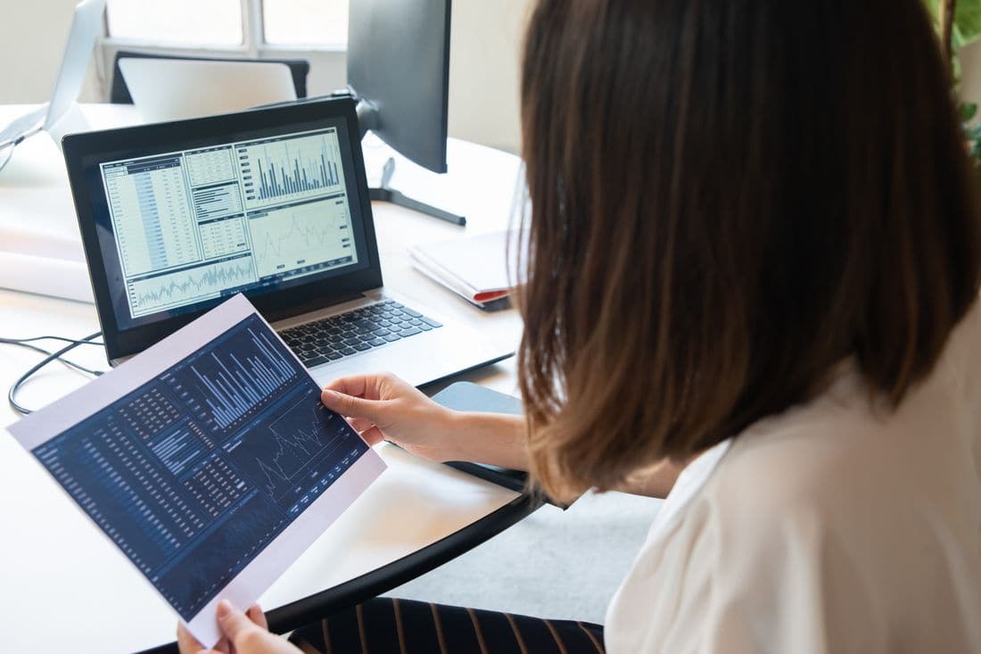 A woman holding a printed page of charts while looking at a laptop screen full of tables and graphs