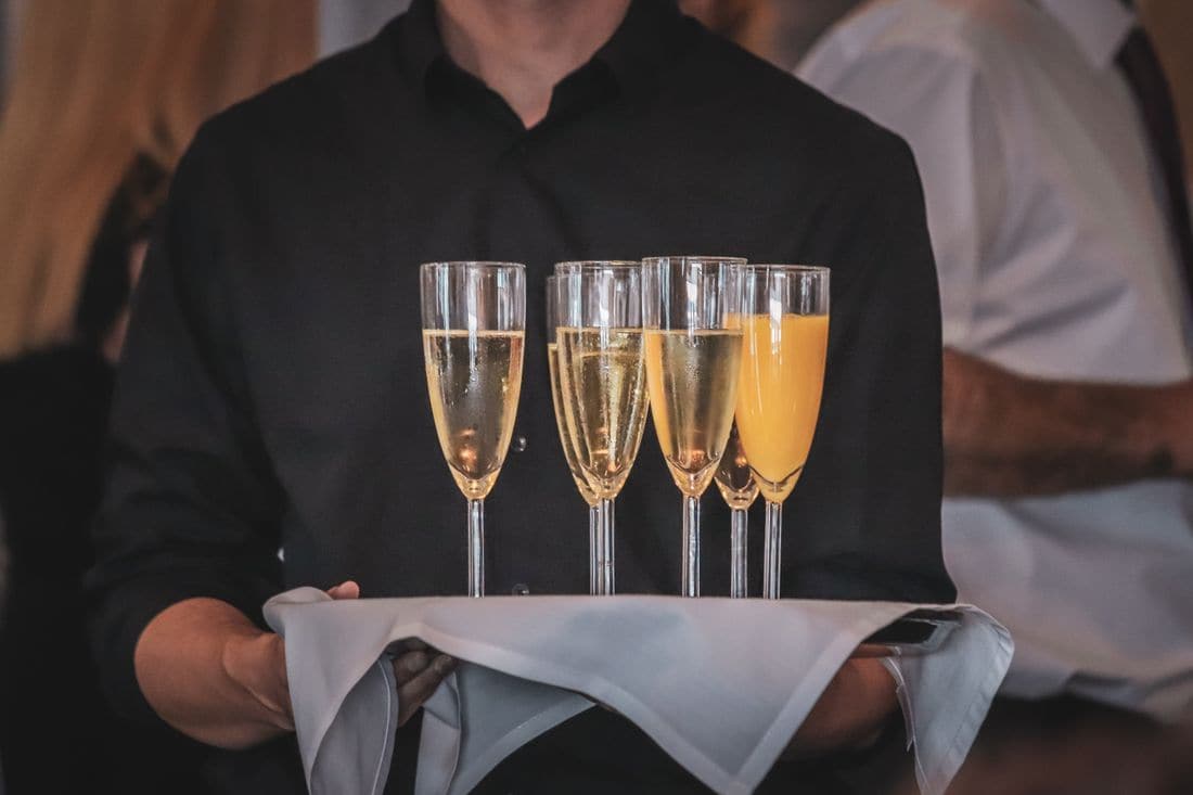 A waiter in a black shirt carrying a tray of champagne and orange juice glasses on a white cloth at an event