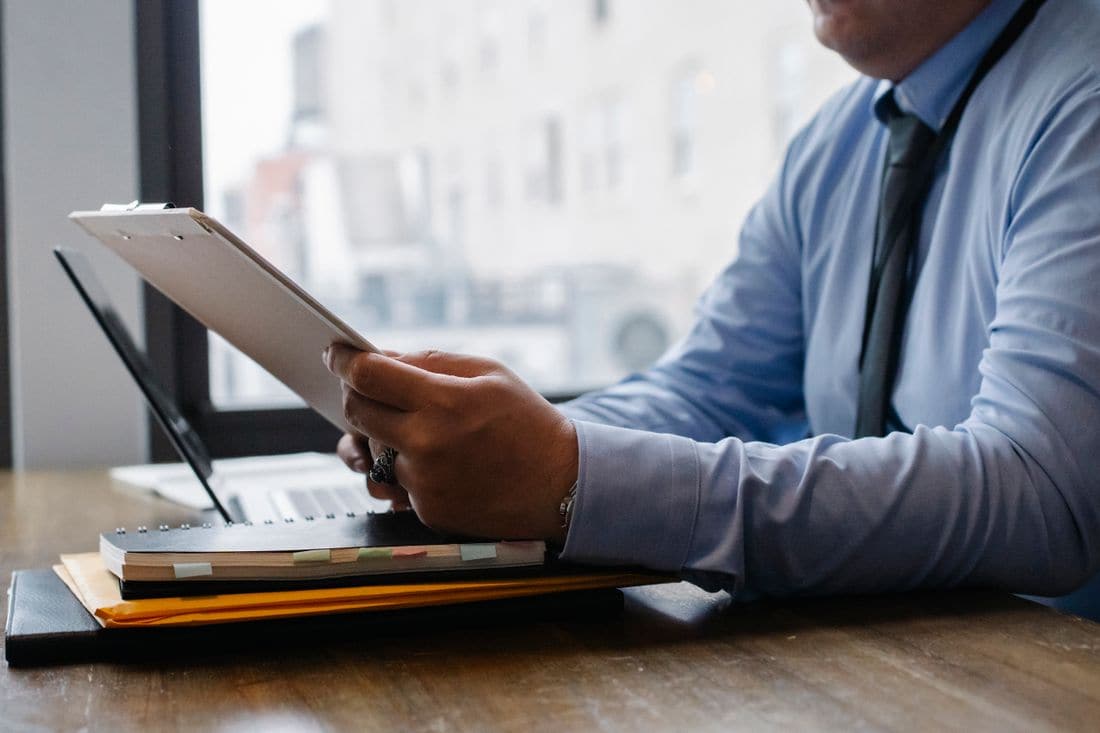 A man in a shirt and tie reading a clipboard at a desk beside a laptop and a stack of notebooks