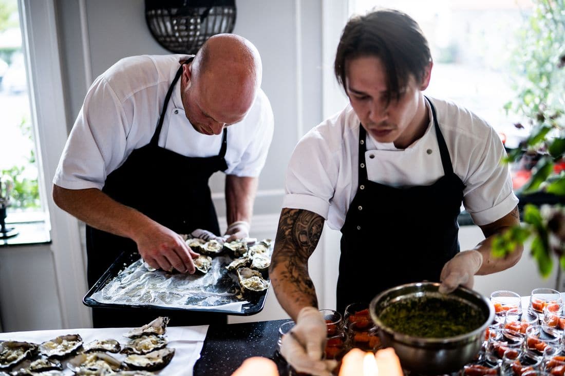 Two chefs in white shirts and black aprons working side by side at one table, one laying out oysters on a tray