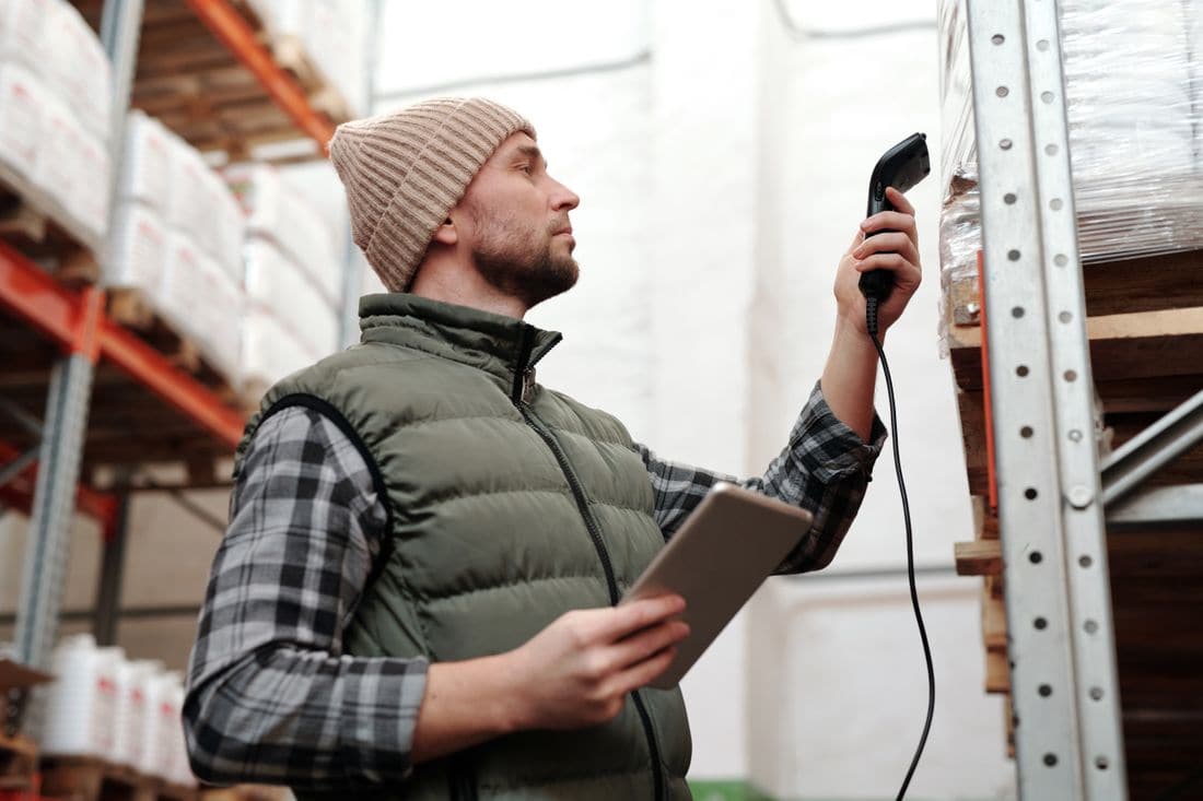 A man in a knitted beanie and padded vest holding a tablet and pointing a handheld barcode scanner at stock on warehouse shelving