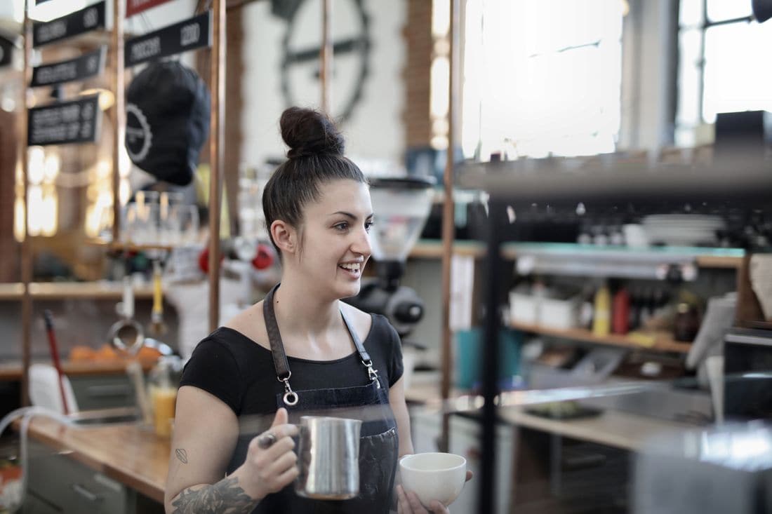 A smiling young barista in an apron holding a milk jug and a cup behind a cafe counter