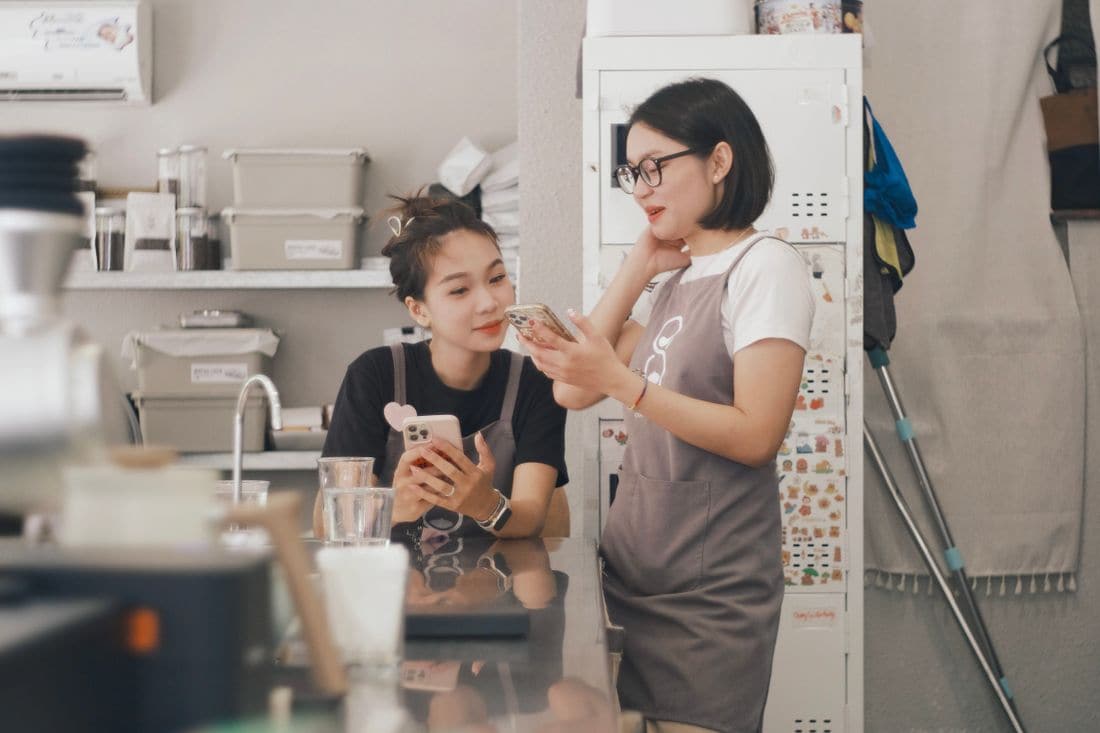 Two cafe workers in aprons looking at a phone together behind the counter