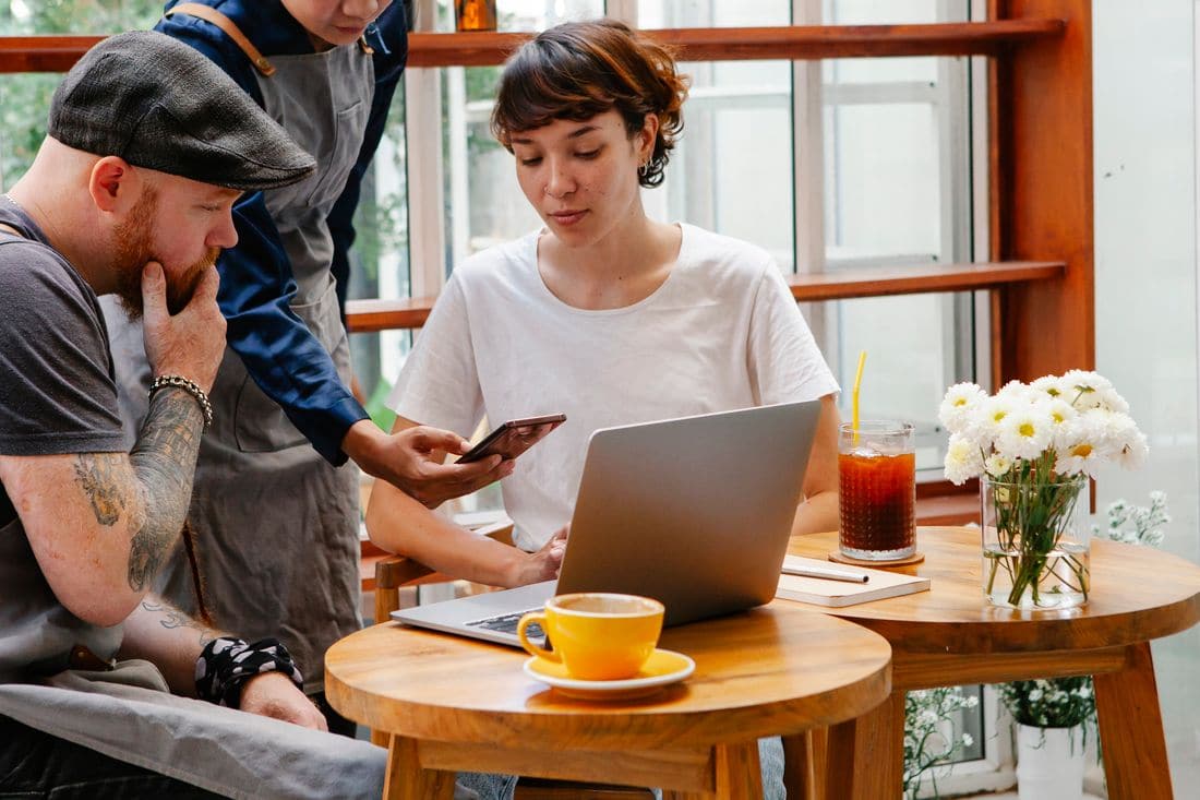 A worker in an apron showing a phone to two people at a cafe table with a laptop and a cup of coffee