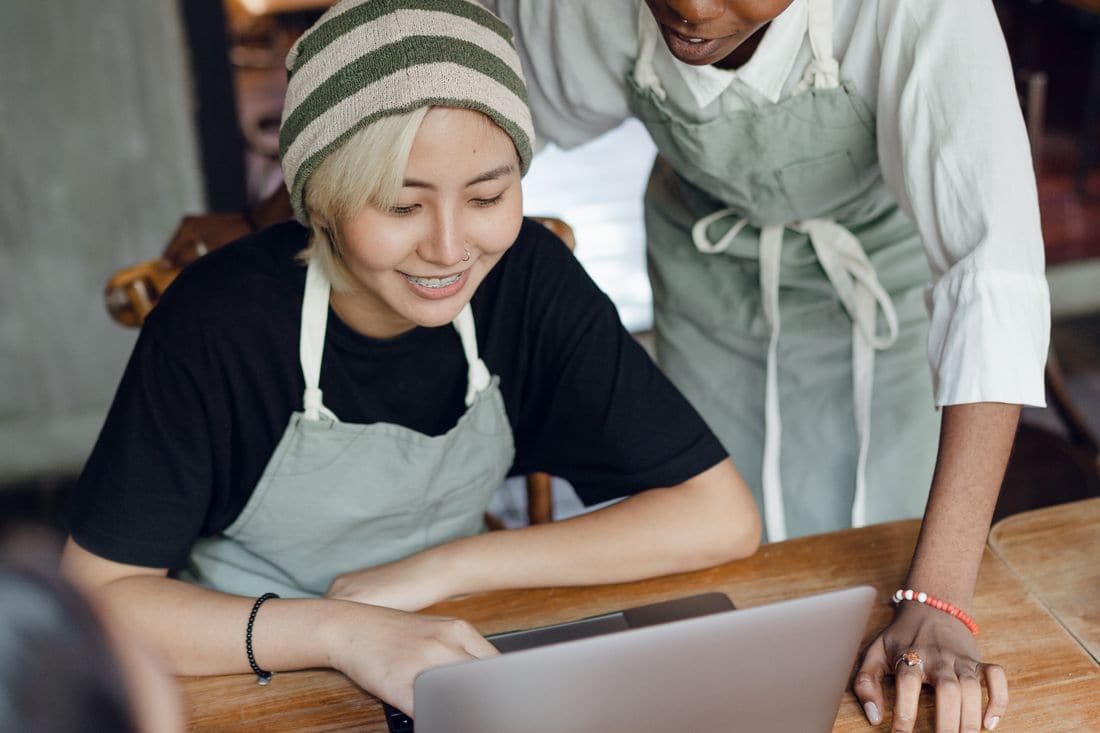 Two cafe workers in aprons looking at a laptop together at a wooden table