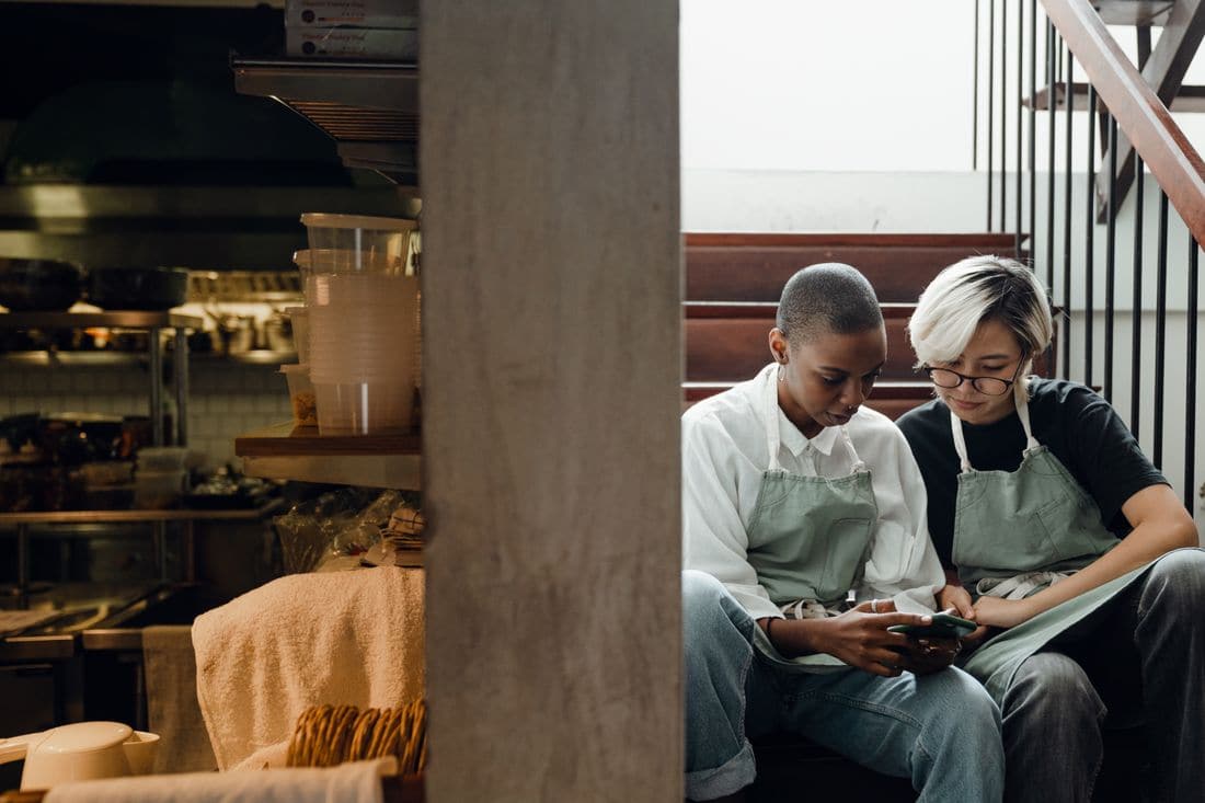 Two kitchen workers in aprons sitting together on the back stairs, checking a phone