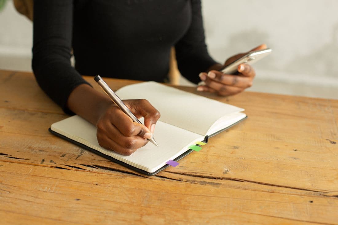 A woman in a black top writing in an open notebook at a wooden table while holding a phone in her other hand
