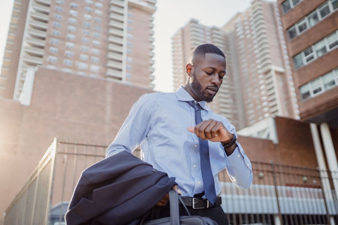 A man in a blue shirt and tie checking his watch as he walks outside with a jacket and bag, apartment towers behind him