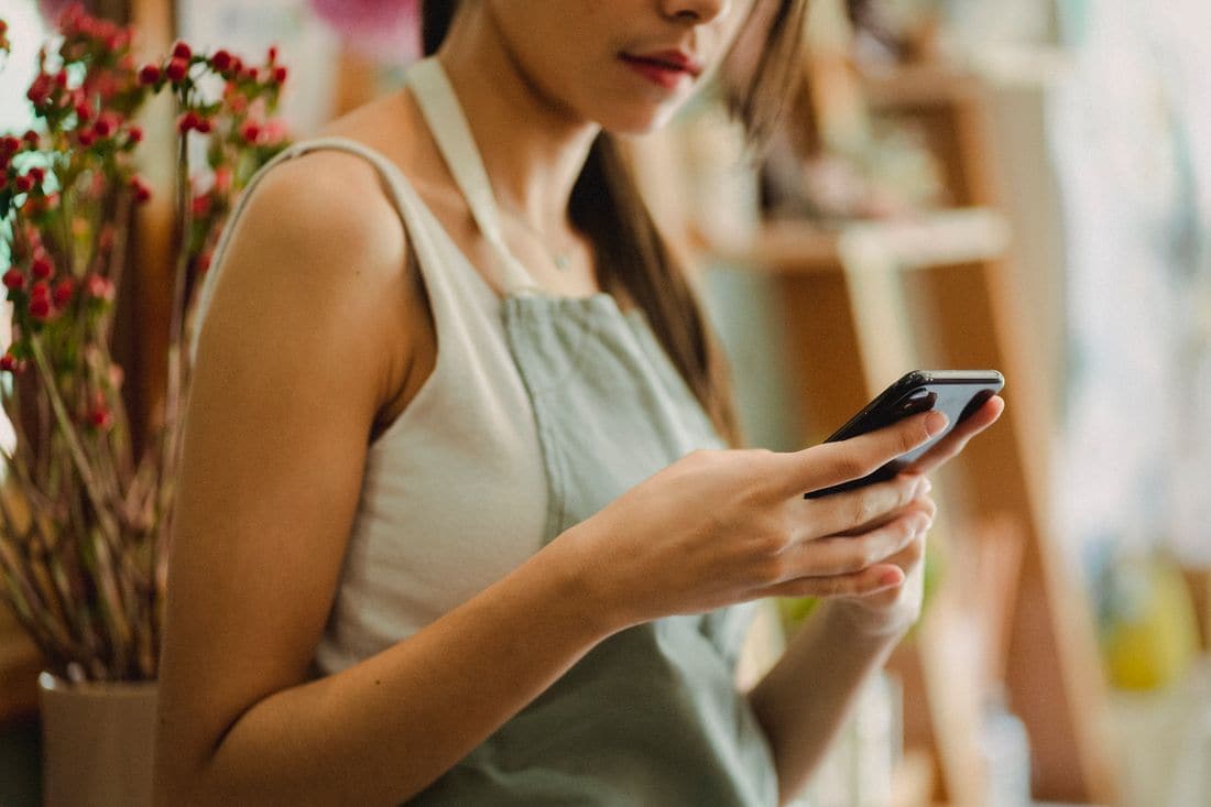 A worker in a grey apron looking at a smartphone in a shop with shelves and red berries behind her