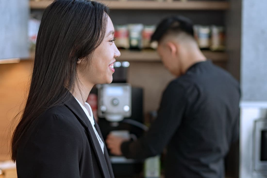 A smiling woman in a black jacket waiting while a coworker uses the coffee machine in a staff kitchen