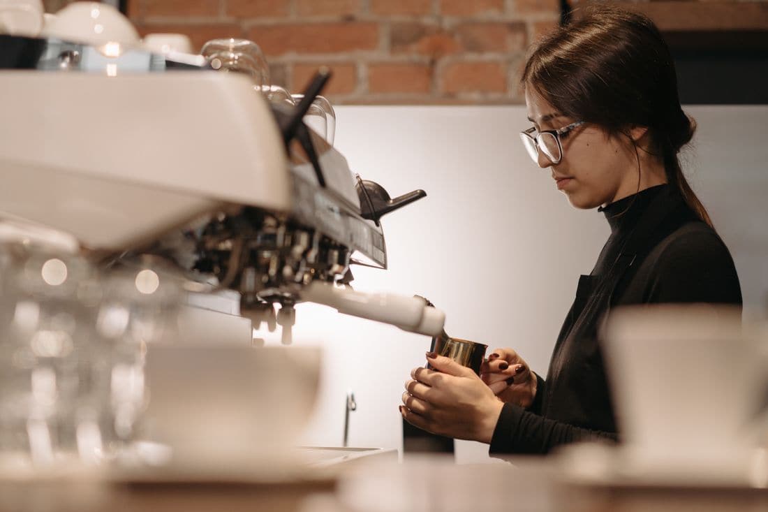 A barista in glasses and a black apron steaming milk in a metal jug at an espresso machine